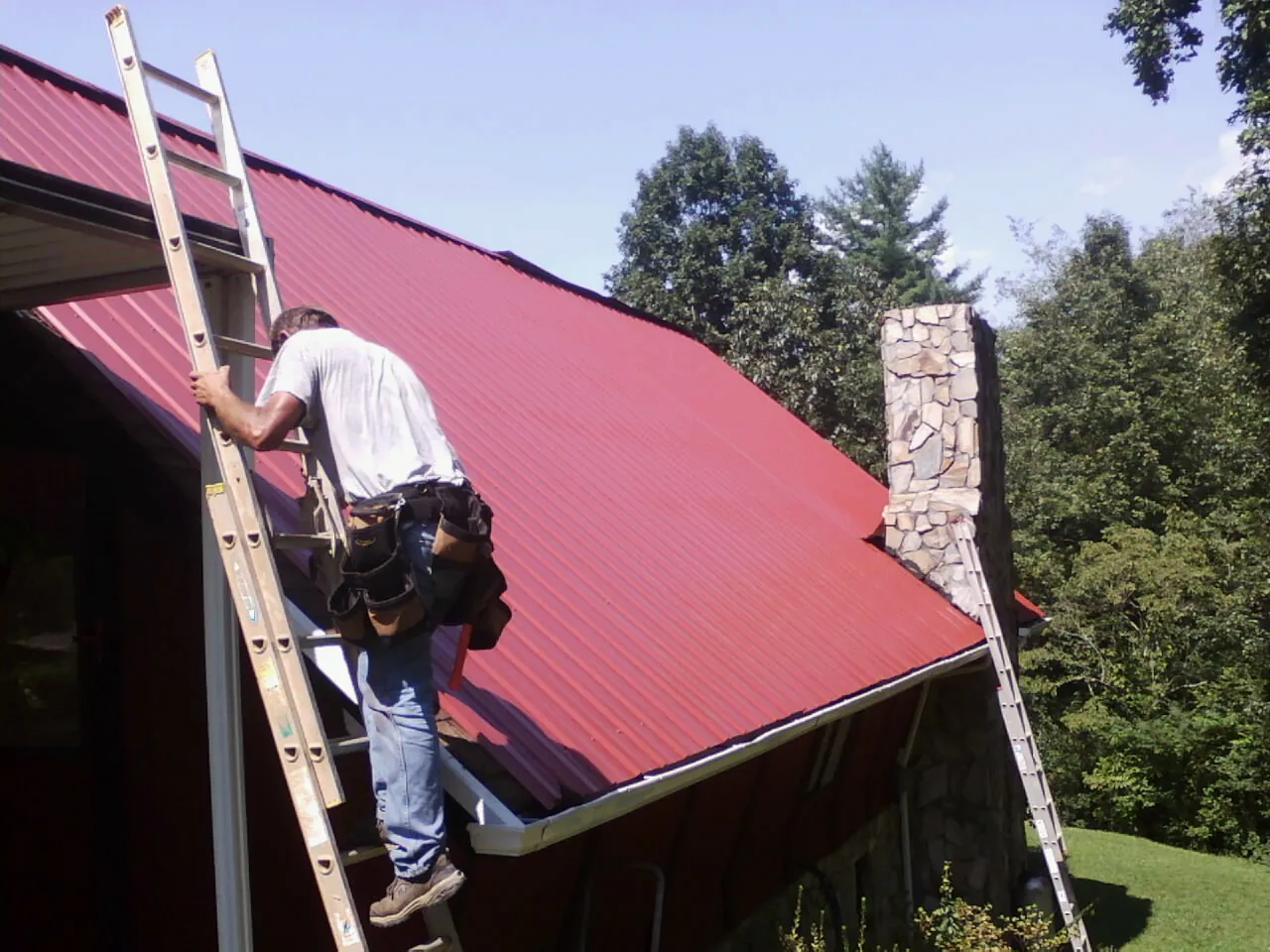 Roofer on a ladder beside a red metal roof and stone chimney — Soffit & Fascia Repair by Roofing Brothers in Chocorua